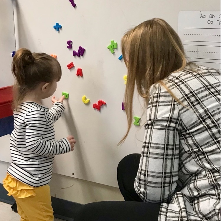 Teacher watches on as student  puts magnet letters on a board