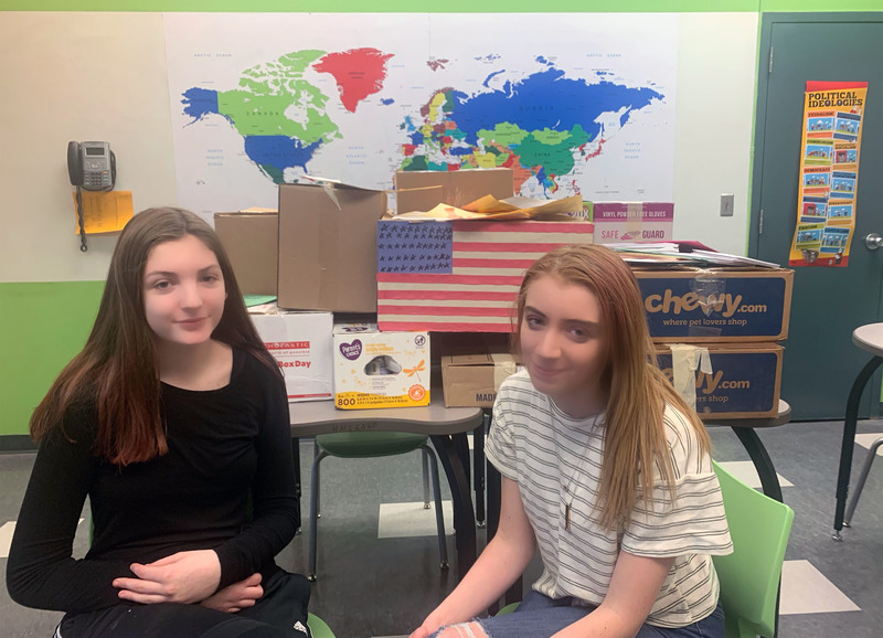 Two students sitting in front of boxes of donations