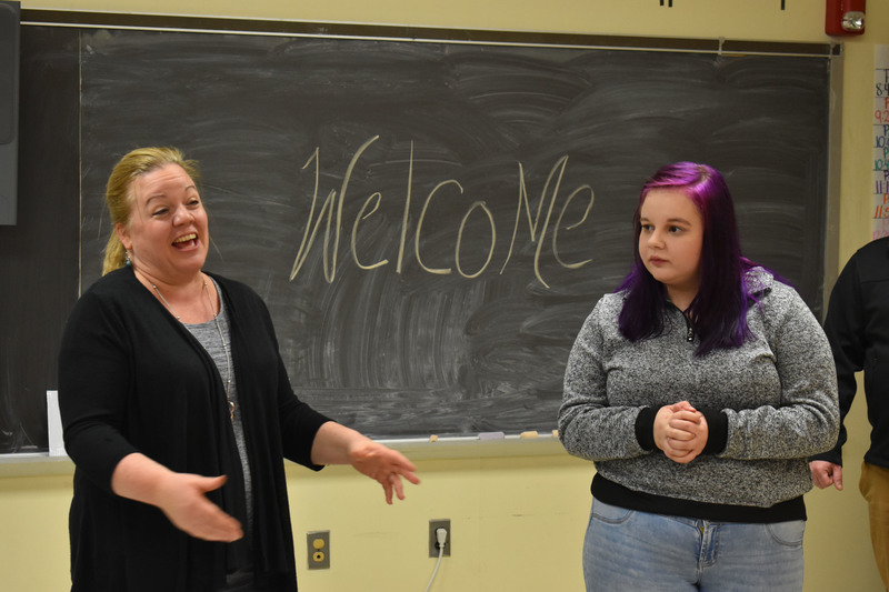 Student and teacher talking to tour guests with a chalkboard behind them with the word Welcome written on it 
