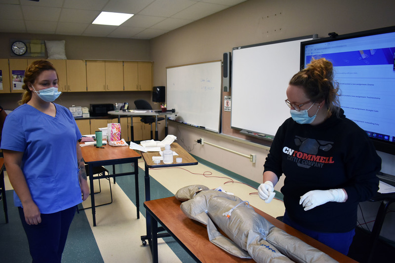 One nursing student in a mask practices on a mannequin on a desk in a classroom while another nursing student in a mask looks on