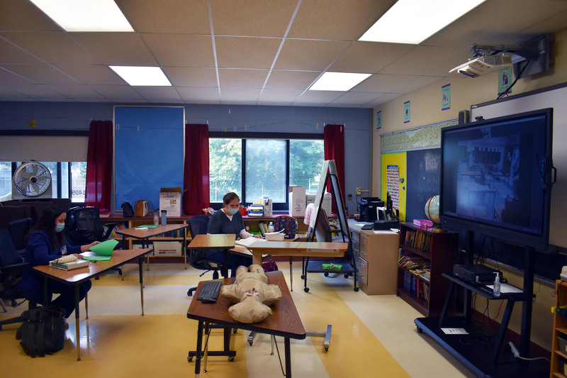 Two nursing students in masks sit at their desks looking at books. There also is a mannequin on the desk and large screen for virtual learning.