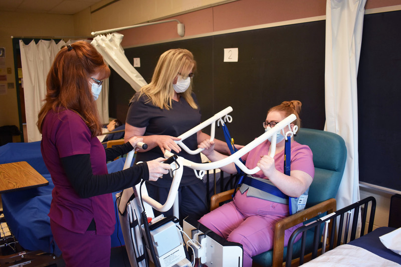 Two adult nursing students use a sit-to-stand lift on another student