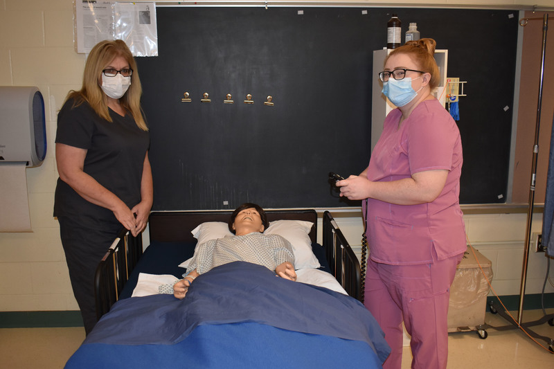 Two adult LPN students pose by a new bed purchased using a grant