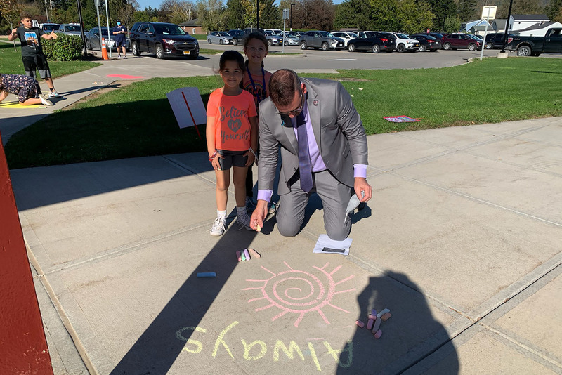 Frankfort-Schuyler Superintendent kneels next to two elementary students and draws with chalk