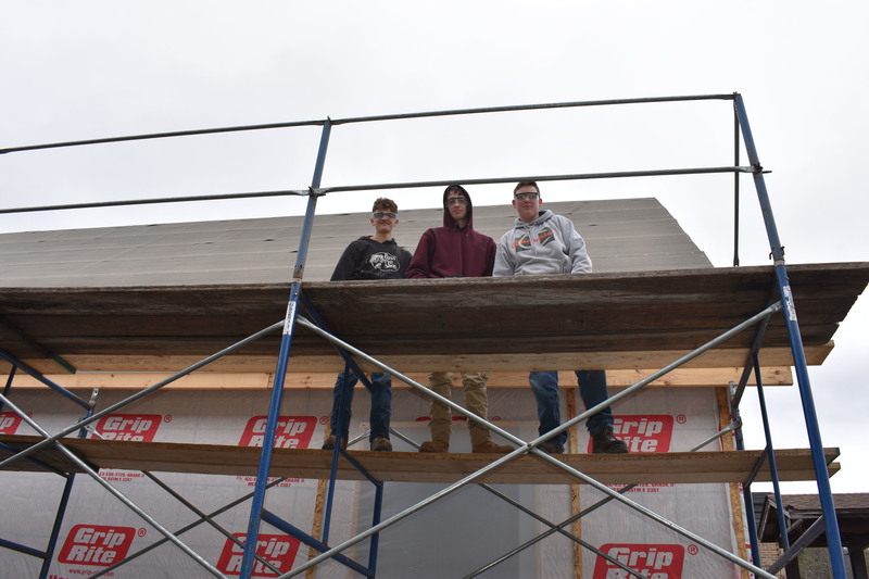 Three students stop to pose for a photo while working on the roof of a house