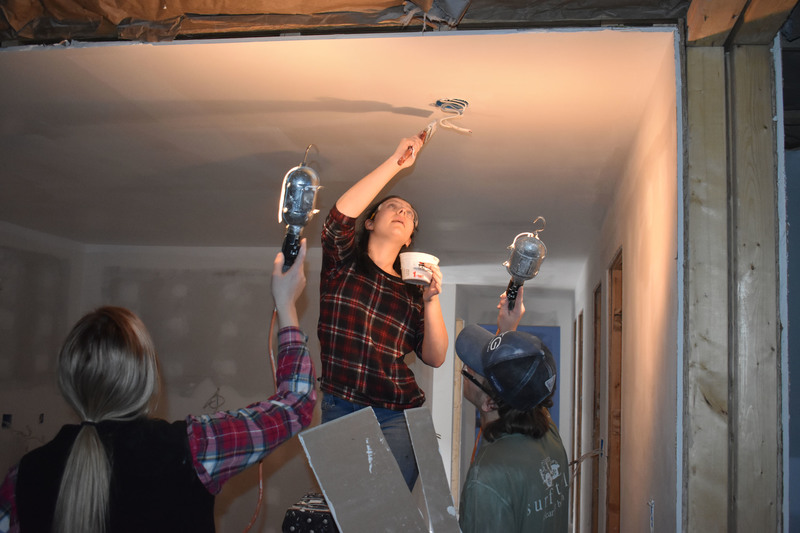 Student on a ladder paints ceiling as others hold up lights