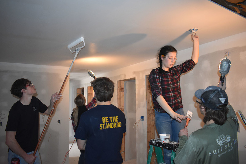 Two students painting the ceiling as other students and teaching assistant hold up lights