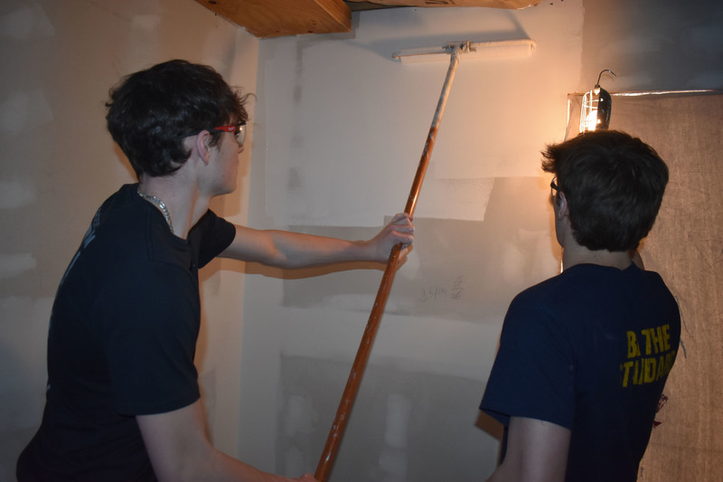 Student paints the wall of a house as another student holds up a light