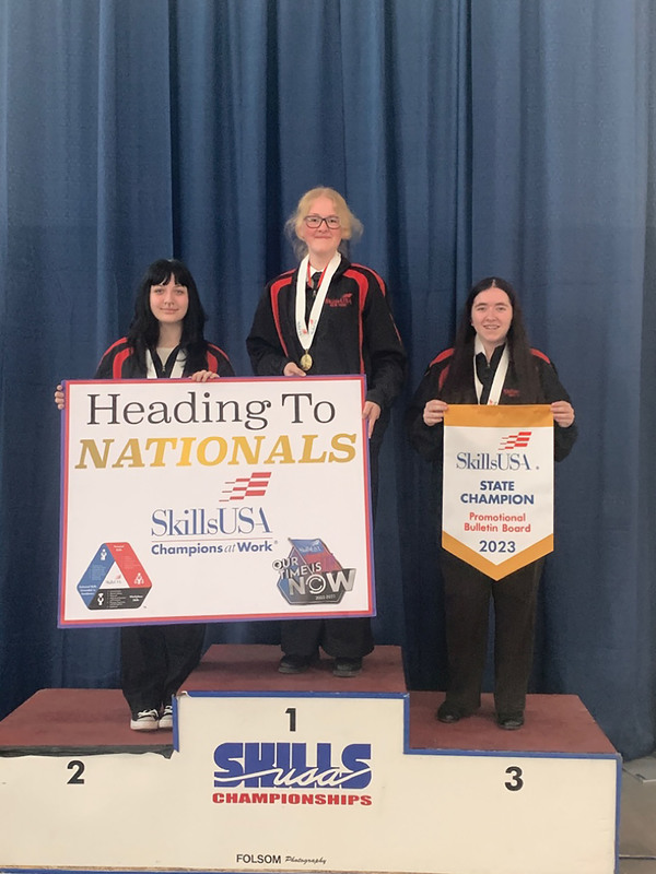 Three Visual Communications Media Arts students holding banners that they won the SkillsUSA state championship and the heading to nationals banner